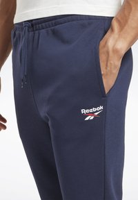 Mørkeblå træningsbukser lavet af blødt stof, med elastisk talje og snøre samt sidelommer, med et Reebok-logo på låret.