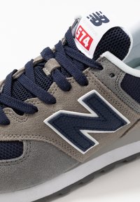 Baskets grises et bleu marine avec des matériaux en daim et en maille. Présentent des lacets épais et un grand logo "N" sur le côté. Marque emblématique New Balance sur la languette.