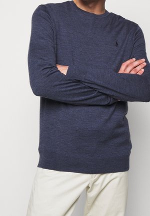 Mann mit verschränkten Armen, der einen dunkelblauen, langärmligen Pullover und hellbeige Hosen vor einem einfarbigen Hintergrund trägt.