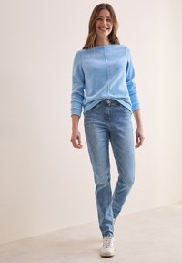 Jasnoniebieski sweter o teksturowym wykończeniu, okrągłym dekolcie i ściągaczach, zestawiony z wyblakłymi niebieskimi dżinsami i białymi trampkami.