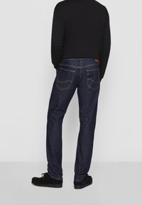 Vaqueros de mezclilla azul oscuro con un corte recto, con dos bolsillos traseros con detalles de costura, combinados con una camiseta de manga larga negra y zapatos negros.