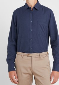 Man med en marinblå skjorta med knappar och bröstficka, samt beiga byxor med knäppt midjeband.