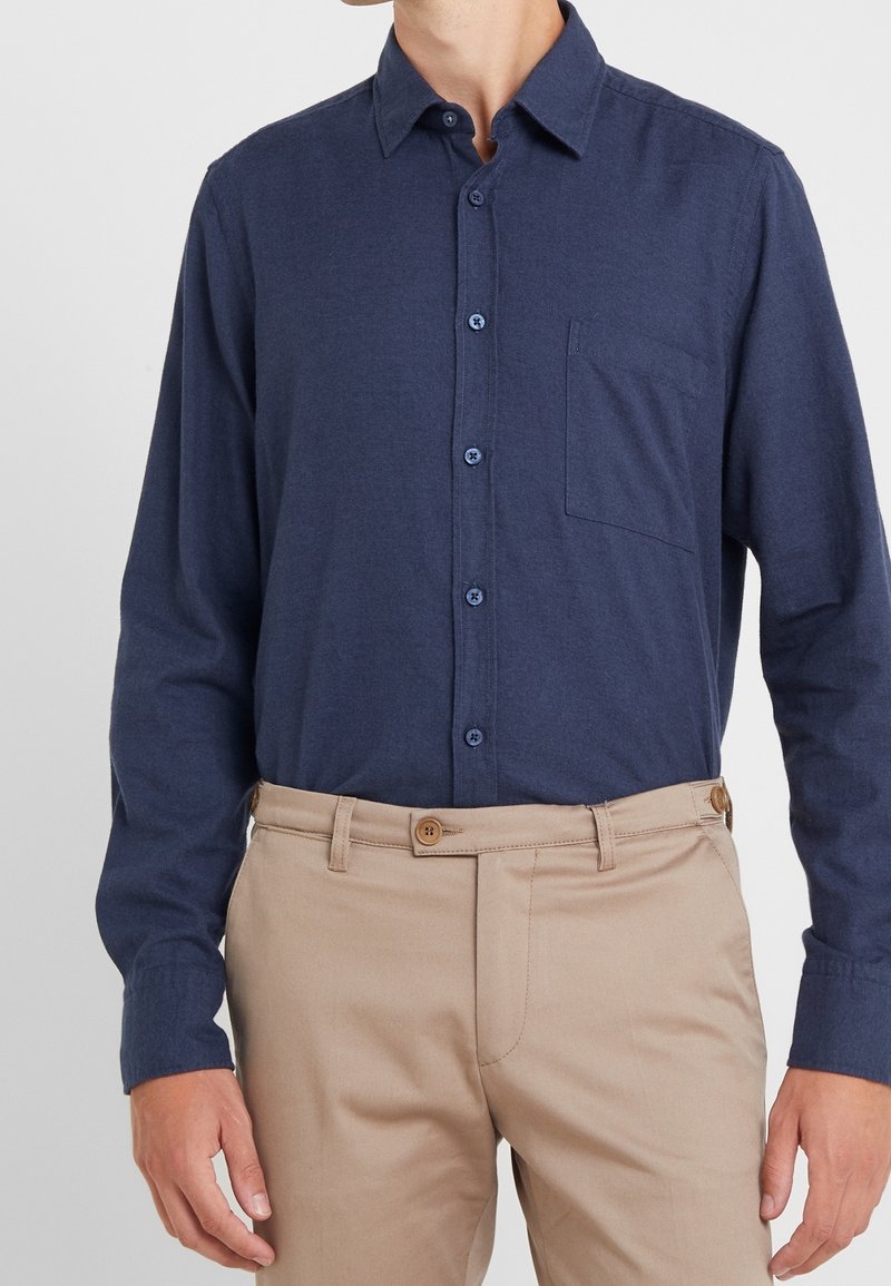 Man med en marinblå skjorta med knappar och bröstficka, samt beiga byxor med knäppt midjeband.