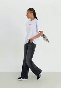 Tricou alb din bumbac cu text roșu, asortat cu blugi negri evazați și adidași negri cu dungi albe. Modelul stă în profil.