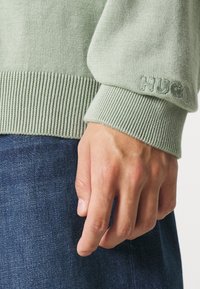 Lichtgroene gebreide trui met geribde manchetten, met "HUGO" geborduurd op de mouw. Denim jeans zijn gedeeltelijk zichtbaar.