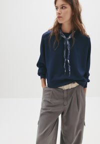 Felpa blu navy con collo a giro e vestibilità ampia, abbinata a pantaloni cargo grigi chiari e una cintura beige, con un foulard a motivo annodato.