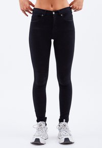 Schwarze Slim-Fit-Jeans mit einer glatten Textur, ausgestattet mit einem Knopfverschluss und fünf Taschen. Kombiniert mit weißen Sportschuhen.