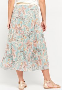 Floral-printed, pleated maxi skirt in light blue with paisley pattern. Features a gold elastic waistband and flowy fabric. Barefoot sandals visible.