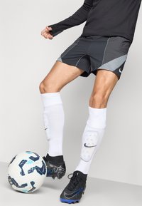 Svart långärmad topp, grå och svarta shorts med meshdetaljer, vita knähöga strumpor, svarta fotbollsskor med blå accenter och en mönstrad fotboll.