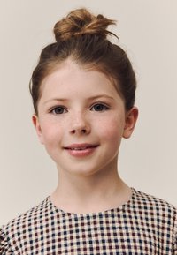 Dievča so svetlohnedými vlasmi zaviazanými do vrkoča na temene hlavy, oblečená v károvaných hnedo-krémových šatách s krátkymi nafúknutými rukávmi, jemne sa usmievajúca.