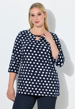 Blondynka w granatowej tunice w białe groszki z rękawami do łokcia, pozująca na białym tle.