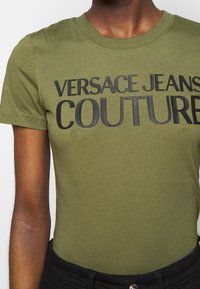 Olivově zelené bavlněné tričko s výrazným černým nápisem "VERSACE JEANS COUTURE". Krátké rukávy a kulatý výstřih.