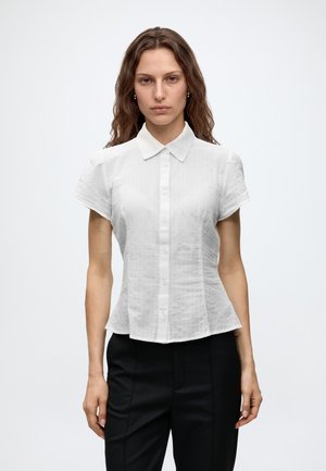 Woman with shoulder-length wavy hair wearing a short-sleeve white button-up shirt and black pants, standing against a plain white background.