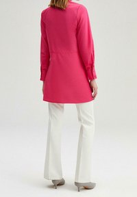 Camisa larga rosa con cuello, mangas abotonadas y corte entallado, combinada con pantalones acampanados blancos y zapatos de tacón grises.