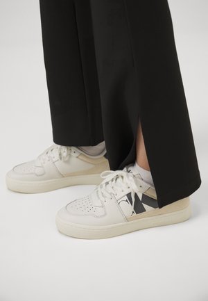 Osoba nosząca czarne spodnie o szerokich nogawkach oraz białe skórzane sneakersy z beżowymi i czarnymi akcentami, stojąca na białej powierzchni.