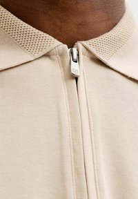 Col en tissu beige tricoté avec fermeture éclair sur une chemise, présentant un détail texturé du col sur une peau foncée.