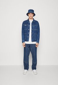 Completo di giacca e pantaloni in denim di colore blu scuro, dotato di numerose tasche, apertura con bottoni e cuciture a contrasto. Abbinato a un cappello a secchiello blu.
