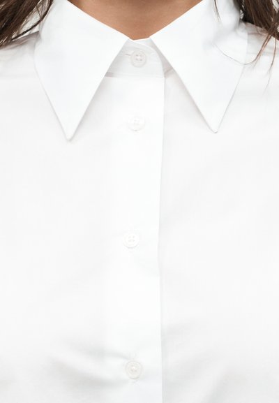 Κοντινό πλάνο ενός ατόμου που φοράει λευκό πουκάμισο με κουμπιά και μυτερό γιακά, με σκούρα μαλλιά ορατά στις άκρες.