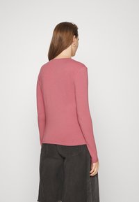 Långärmad, figurnära t-shirt i dammig rosenfärg, tillverkad av ett mjukt material med rund halsringning och en böjd nederkant, tillsammans med mörka denimbyxor.