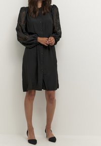 Woman in a black buttoned dress with sheer lace puff sleeves and black high-heeled shoes standing against a plain background.