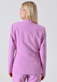 Vrouw met blond haar in een op maat gemaakt lila colbert en bijpassende broek, met de rug naar de camera gekeerd tegen een effen lichte achtergrond.