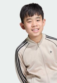 Giacca beige con zip e strisce nere lungo le maniche, con un piccolo logo nero sul petto, realizzata in tessuto liscio.