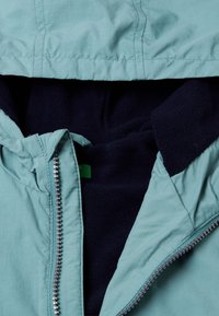 Světle modrý kabát s měkkým námořnickým fleecem uvnitř. Obsahuje zip a štítek. Tkanina je hladká s viditelnými detaily šití.