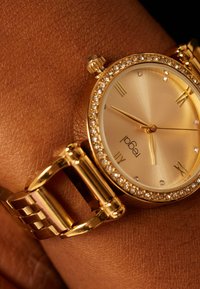 Goldene Armbanduhr mit Strassstein-Lünette, römischen Ziffern bei 12, 3, 6, 9 und goldenem Armband auf dunkelbrauner Handgelenkshaut.