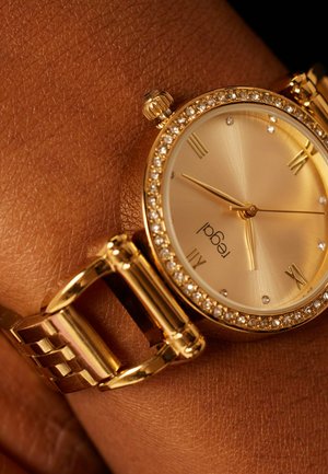 Gouden polshorloge met strassteen lunette, Romeinse cijfers op 12, 3, 6, 9 en gouden armband aan een donkerbruine polshuid.