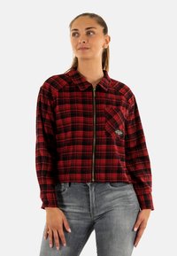 Donna che indossa una giacca a zip rossa e nera a quadri con tasca frontale, abbinata a jeans grigi sbiaditi, in piedi di fronte a uno sfondo bianco.