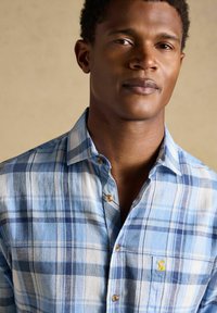 Mies, joka käyttää vaaleansinivalkoista ruudullista napillista paitaa, jossa on pieni keltainen kirjailtu logo rintataskussa, beigeä taustaa vasten.