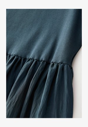 Temno turkizna obleka z ribanim vrhom in razširjenim, nabranim dnom, izdelana iz lahkega, teksturiranega blaga.