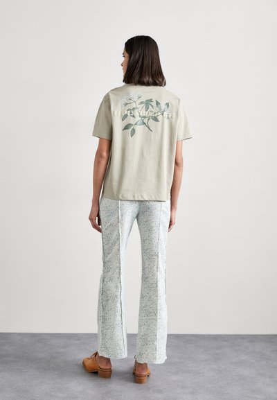 Olijfgroene katoenen t-shirt met botanische print op de achterkant, gecombineerd met lichtgebloemde broeken. Model draagt bruine houten klompen.