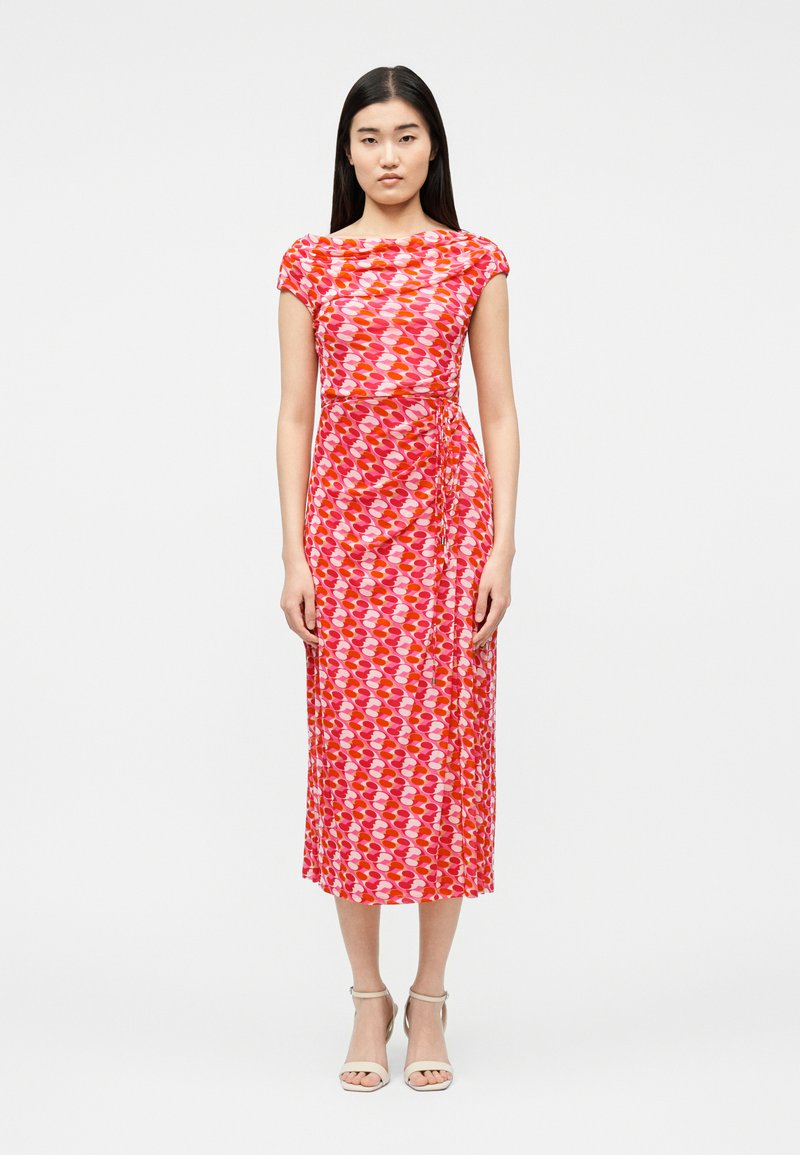 Γυναίκα που στέκεται φορώντας midi φόρεμα με κόκκινο και ροζ μοτίβο, με κοντά μανίκια τύπου καπέλου και λευκά πέδιλα με ανοιχτά δάχτυλα, σε απλό φωτεινό φόντο.