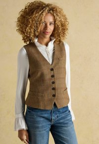 Hnedý károvaný sveter s piatimi gombíkmi, dvoma prednými vreckami, skombinovaný s bielou košeľou s golierom a džínsami. Mäkka textúra látky.