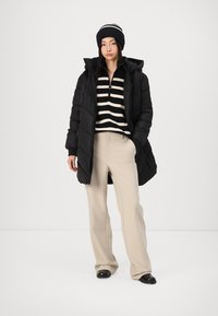 Doudoune noire avec design matelassé, portée par-dessus un haut rayé noir et beige. Associée à un pantalon beige et à un bonnet rayé noir.