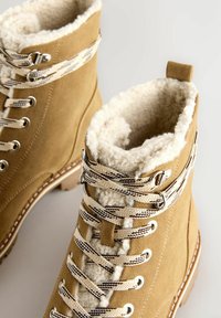 Bruna mockakängor för vintern med vit fleecefoder och beige snören i vandringsstil med svarta ränder på en ljus bakgrund.