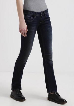 Kobieta ubrana w ciemnoniebieskie dopasowane dżinsy, szarą bluzkę i czarne sznurowane buty, stojąca na tle jednolitego jasnego tła.