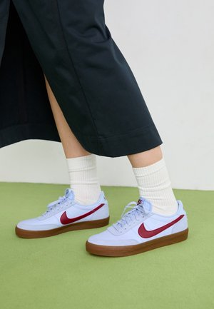 Jambes portant des chaussettes blanches et des baskets blanches Nike avec un swoosh rouge et des semelles en gomme, debout sur un sol vert contre un mur blanc.