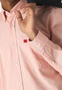 Helle rosa Button-up-Hemden aus einem glatten Stoff, mit einer Brusttasche und einem kleinen roten "HUGO"-Label auf der Brust.