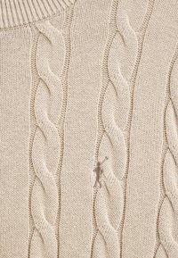 Pull en tricot beige présentant un motif à torsades, un col rond et un petit logo brodé sur le devant.