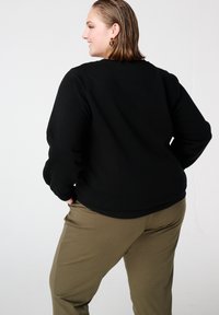 Femme aux cheveux mi-longs portant des boucles d'oreilles en or, un chemisier noir à manches longues et un pantalon olive, légèrement tournée sur le côt�é avec un sourire.