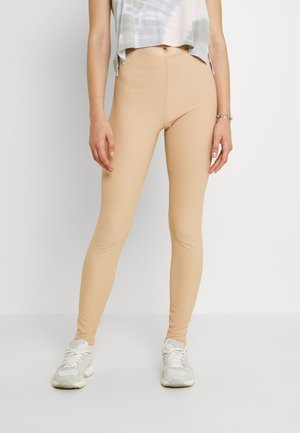 Leggings stretch beiges avec une texture côtelée, taille haute, accompagnés d'un haut court en tie-dye et de chaussures de sport gris clair.