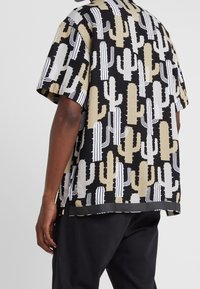 Chemise à manches courtes avec une base noire et un motif de cactus en beige, blanc et noir. Coupe décontractée avec un ourlet arrondi et des fentes sur les côtés.