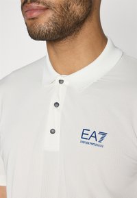 Λευκό πόλο μπλουζάκι με υφασμάτινο σχέδιο, διαθέτει πλάκα με δύο κουμπιά, σκούρα κουμπιά και μπλε λογότυπο EA7 Emporio Armani στο στήθος.