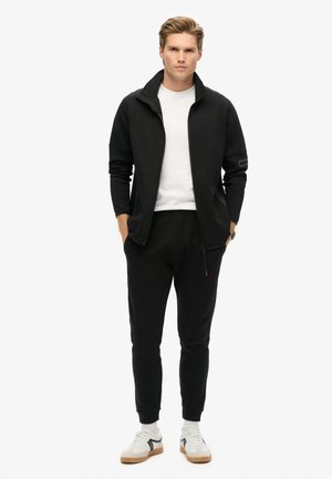 Black zip-up jacket with high collar, paired with matching sweatpants. Model wears a white t-shirt and white sneakers with black accent.