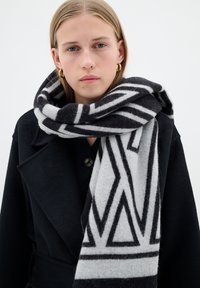 Giovane con lunghi capelli biondi che indossa orecchini a cerchio dorati, un cappotto nero e una sciarpa spessa a motivi geometrici bianco e nero.