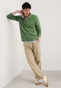 Grüner Sweatshirt mit Viertelreißverschluss und gerippten Bündchen, über einem gestreiften Hemd getragen. Beige lässige Hose und weiße Sneakers mit braunen Sohlen.