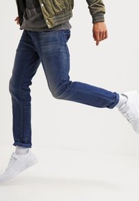 Jeans en denim bleu avec une coupe slim et une légère déchirure, associés à des baskets blanches. Les jeans sont retroussés aux chevilles, laissant voir les chaussures.