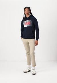 Felpa navy con un audace design grafico che presenta il testo "JACK & JONES" in rosso. Un paio di chinos beige arrotolati e sneakers bianche e nere.